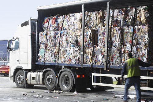 A large commercial waste truck with an open side panel, filled with tightly compressed bales of mixed recyclable rubbish, primarily paper and cardboard. The bales exhibit various colours and textures including browns, whites, and slight touches of blue and green, reflecting different types of used packaging materials. The truck is parked on a paved surface, likely in a commercial or industrial area, with some waste debris scattered on the ground nearby. To the right, a person wearing high-visibility clothing and jeans is walking past, possibly a waste management worker. The background features a clear sky and hints of other vehicles or structures typical of a waste collection or disposal site in Kenton. The scene aligns with waste removal services provided by Commercial Waste Kenton, emphasizing efficient handling of bulky rubbish in a professional environment.