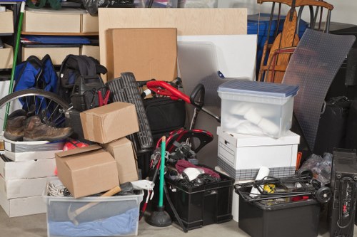 A cluttered indoor space, possibly a garage or storage area, filled with various household items awaiting rubbish removal. Visible in the scene are multiple cardboard boxes of different sizes, some sealed and others partially open, stacked on top of each other. There are clear plastic storage containers, one with a grey lid containing what appears to be soft fabric or clothing, and another beneath it holding miscellaneous items. A black toolbox and a few smaller black plastic crates are also present, holding tools and parts. Leaning against the boxes is a red and black manual hand cart, and nearby, a green-handled mop is propped against a box. In the background, a metal shelving unit holds additional boxes and miscellaneous objects, and parts of chairs and other furniture can be seen, hinting at a mix of household and possibly garden items. The environment has a utilitarian, slightly cluttered appearance typical of a space scheduled for waste clearance by Commercial Waste Kenton, located in the London Borough of Brent, with the scene lit by ambient lighting that highlights the assortment of materials and textures, including corrugated cardboard, plastic, metal, and fabric.