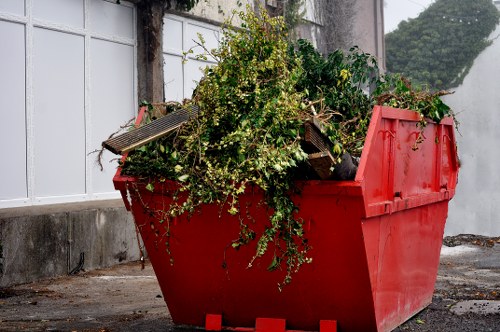 A large red skip filled with gardening waste, including green branches, small leafy shrubs, and overgrown plant material, positioned on a paved driveway. The waste appears freshly collected, with some loose leaves and tendrils spilling over the edges of the skip. Surrounding the skip, there is a concrete wall and a white-framed window with frosted glass behind, indicating a residential or commercial property in Kenton. The environment looks like an outdoor area suitable for rubbish collection, with natural daylight illuminating the scene. The skip, used by Commercial Waste Kenton, is intended for garden and general waste removal, aligning with their services in the local postcode area. The photograph captures the practical aspect of rubbish clearance services in the context of garden or yard clean-up, emphasizing the volume and type of waste that might be handled during a typical waste removal operation in Kenton.