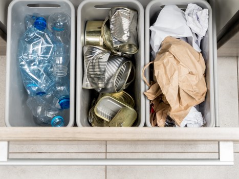 The image displays three grey plastic bins arranged side by side, situated on a tiled floor in an indoor or outdoor setting. The left bin contains several transparent plastic bottles, some crumpled, with blue caps visible on a few. The middle bin holds a variety of metallic cans, predominantly aluminium, some flattened and stacked loosely, with rims and shiny surfaces reflecting ambient light. The right bin contains crumpled paper or paper bags, primarily in beige or off-white tones, with some white paper towels or tissues on top, showing a somewhat disorganized arrangement. The background reveals a clean, tiled surface, suggesting a controlled environment such as a kitchen or waste collection point. The scene is well-lit, with no visible obstructions, providing a clear view for waste disposal or recycling purposes in a commercial or residential setting near Kenton, designed to facilitate rubbish removal services by Commercial Waste Kenton.