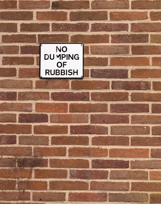 A rectangular metal sign mounted on a red brick wall, with black text on a white background reading 'NO DUMPING OF RUBBISH' in uppercase letters. The sign appears weathered with some minor scratches a
