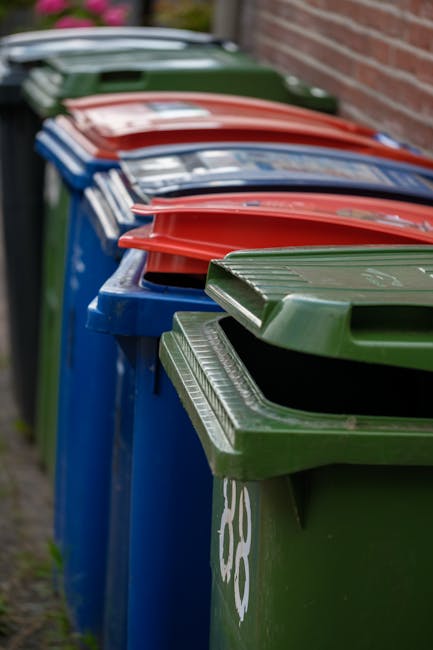 A row of four large wheelie bins positioned on a pavement beside a red brick wall. The bins are blue with textured, ribbed green and red lids, and are made of durable plastic with a smooth, slightly g