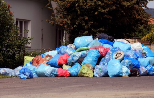 A large pile of discarded rubbish bags, predominantly made of blue plastic, with some in red, green, yellow, and transparent materials, stacked along the edge of a residential street. The bags contain various types of waste and are piled on the pavement close to a white house with a window and a small garden with shrubs and a tree in the background. The scene suggests a waste collection or clearance area, with some bags leaning against a tree and others spread across the driveway and street, possibly awaiting collection by a waste management service such as Commercial Waste Kenton. The environment appears tidy apart from the waste pile, with natural lighting indicating daytime, and the surrounding greenery providing a subtle contact to the local context of Kenton.